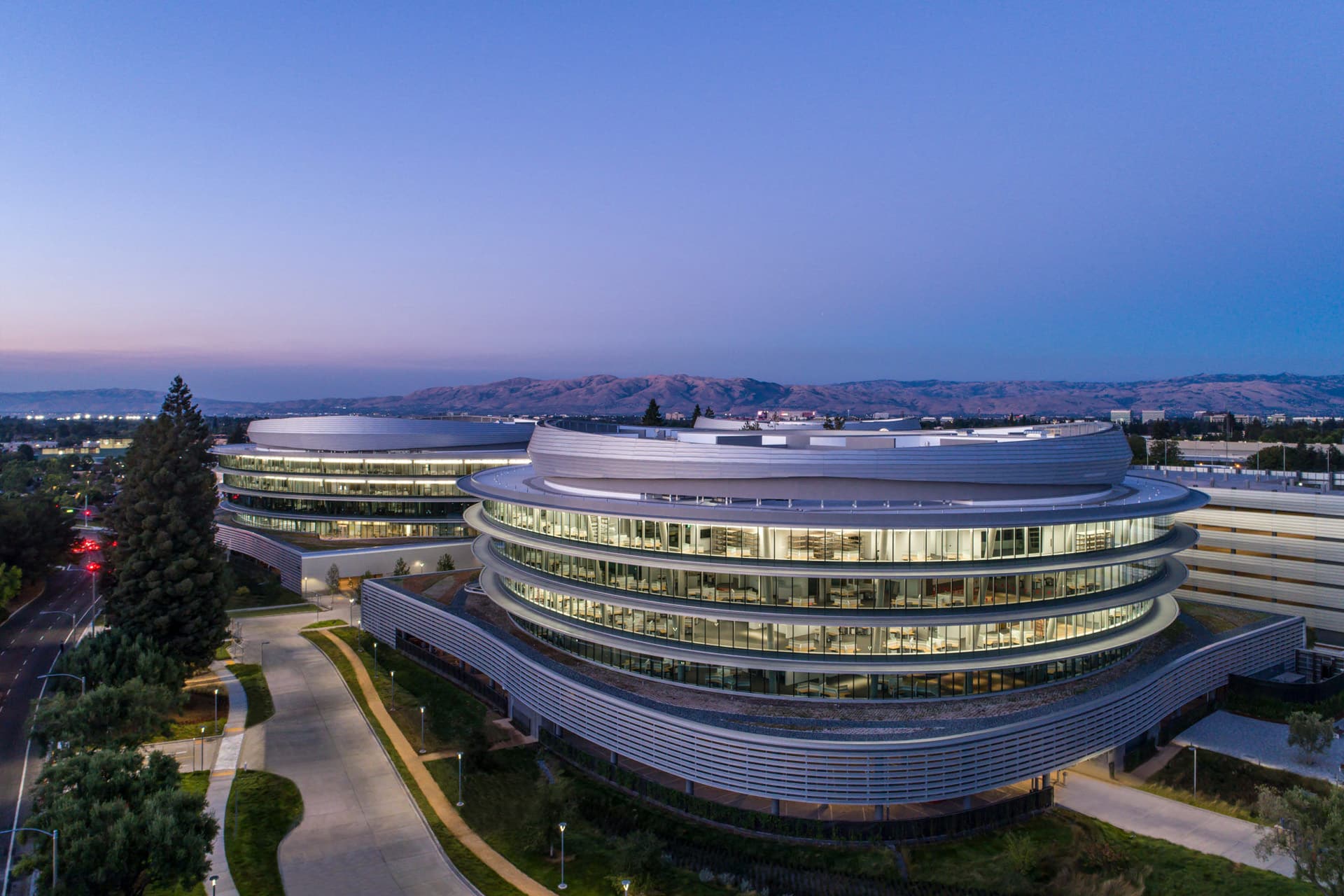 Apple Sunnyvale expansion - Aerial view of a modern, circular office building with glass walls at dusk, surrounded by trees and a distant mountain range under a clear sky.