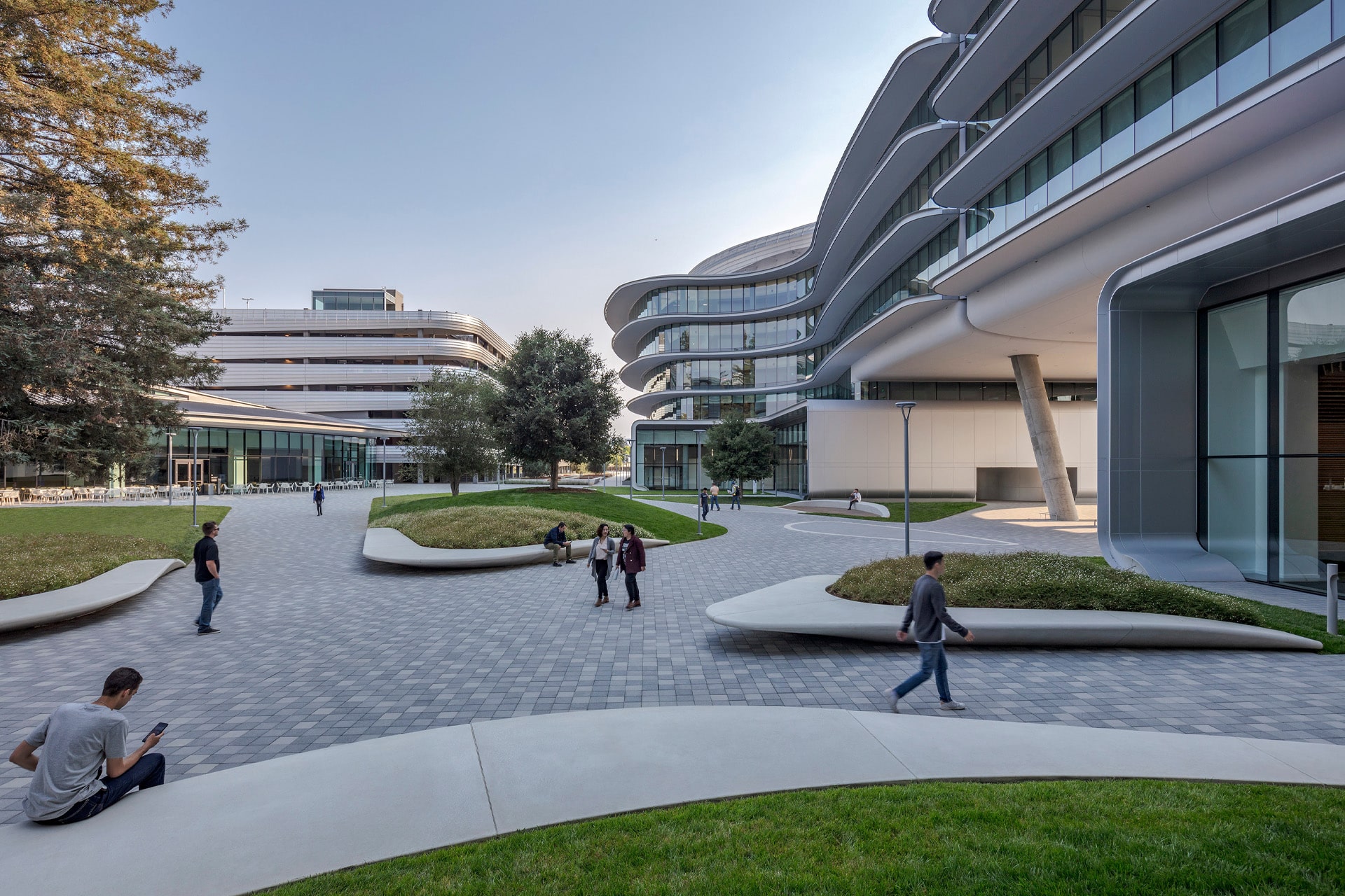 Modern campus courtyard with curved glass buildings, landscaped greenery, paved walkways, and people walking, sitting, and talking outdoors on a clear day.