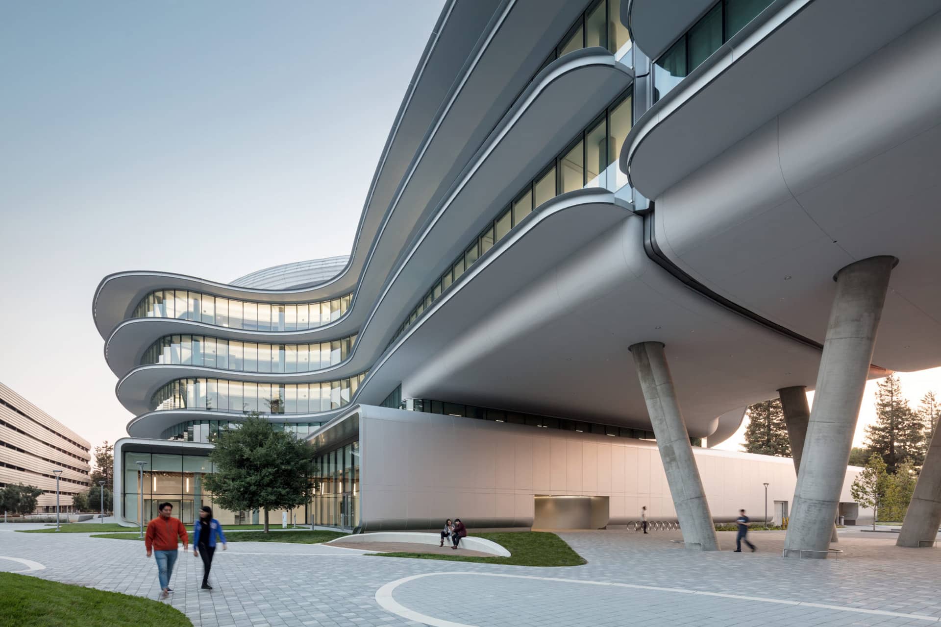A modern, curving glass building with multiple floors, supported by angled columns. People walk and sit on benches along a paved path beside green grass and trees under a clear sky.