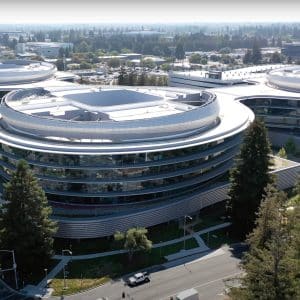Aerial view of a modern circular office building with glass walls and surrounding greenery, intersected by roads and trees, in an urban area on a clear day.