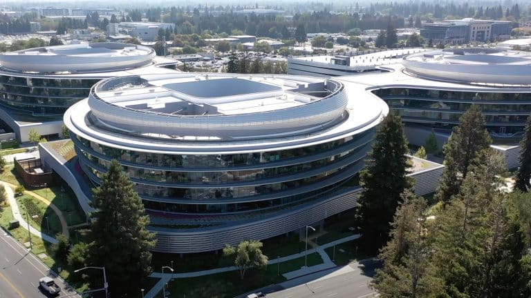 Aerial view of a modern circular office building with glass walls and surrounding greenery, intersected by roads and trees, in an urban area on a clear day.