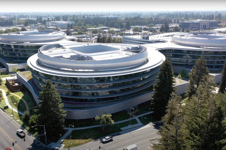 Aerial view of a modern circular office building with glass walls and surrounding greenery, intersected by roads and trees, in an urban area on a clear day.