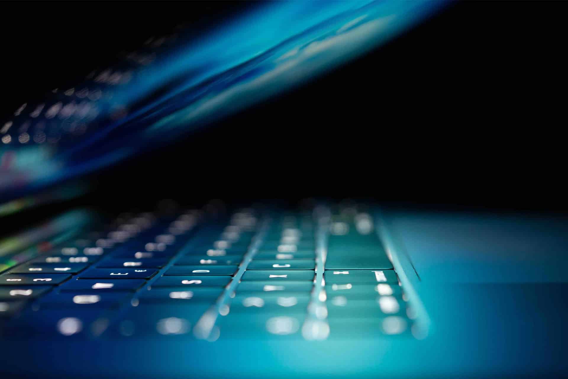 A close-up of a laptop keyboard illuminated by blue light hints at iPhone secrets, with the screen partially closed and casting a soft glow on the keys against a dark background.