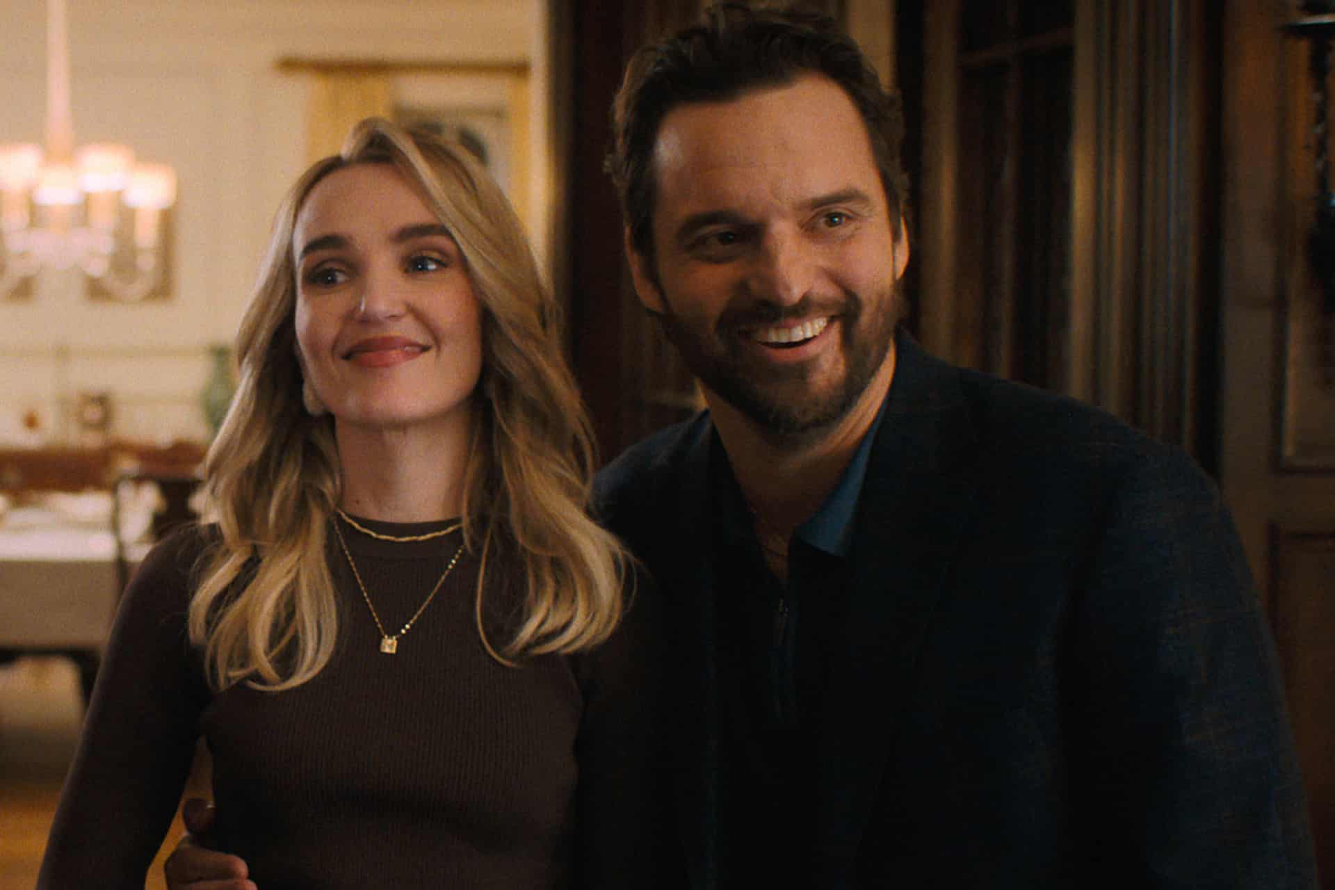 A smiling woman with long blonde hair stands next to a bearded man in a dark jacket. They appear happy and are standing indoors in a warmly lit room, reminiscent of a cozy scene from an Apple TV series.