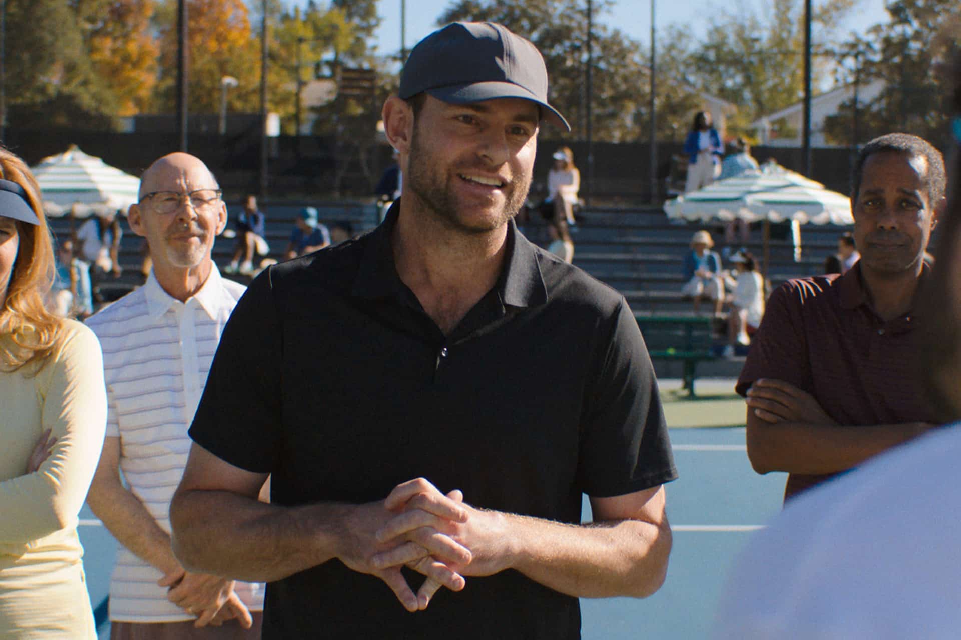 A man in a black shirt and cap smiles while speaking to a group at an outdoor tennis court during The Dink premiere. Several people stand behind him, listening, with trees and umbrellas visible in the background, celebrating the event featured on Apple TV.
