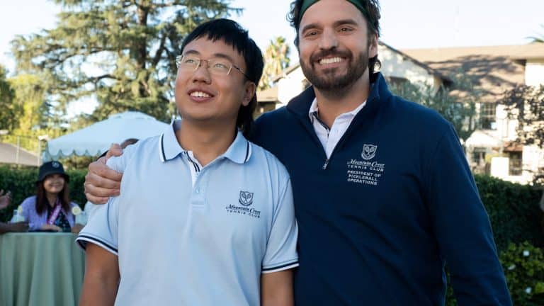 Two men wearing white and navy shirts with embroidered logos stand smiling outdoors, with one arm around each other. Behind them, trees, a lawn, and a person sitting at a table are visible in the background—an atmosphere reminiscent of scenes from an Apple Original Films production featuring stars like Ben Stiller or The Dink.