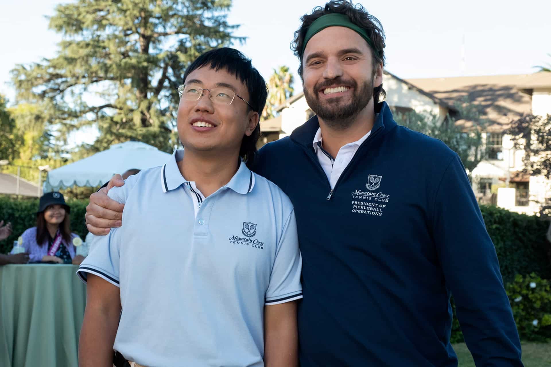 Two men wearing white and navy shirts with embroidered logos stand smiling outdoors, with one arm around each other. Behind them, trees, a lawn, and a person sitting at a table are visible in the background—an atmosphere reminiscent of scenes from an Apple Original Films production featuring stars like Ben Stiller or The Dink.