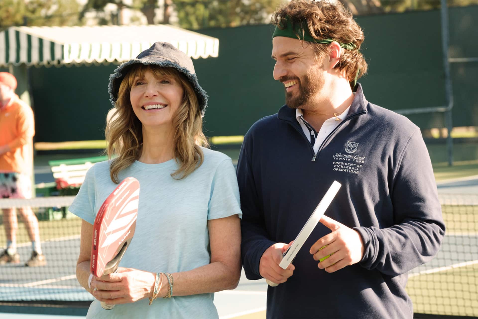Two people stand on a tennis court, smiling and holding pickleball paddles as if ready for a fun match that could belong in an Apple TV scene. The woman wears a sun hat and light blue shirt, while the man—who bears a resemblance to Ben Stiller—sports a dark jacket and headband. There’s a net and green background behind them, evoking the fresh style of an.