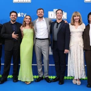 Seven people stand together smiling on a blue carpet in front of a blue backdrop with "Still" written on it, dressed in formal and semi-formal attire, posing for a group photo at The Dink premiere presented by Apple Original Films.