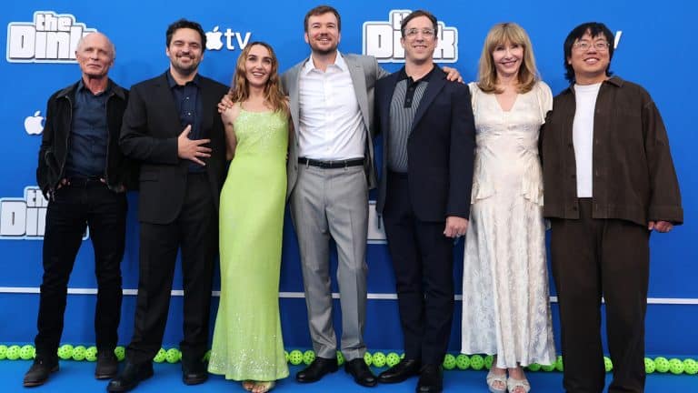 Seven people stand together smiling on a blue carpet in front of a blue backdrop with "Still" written on it, dressed in formal and semi-formal attire, posing for a group photo at The Dink premiere presented by Apple Original Films.
