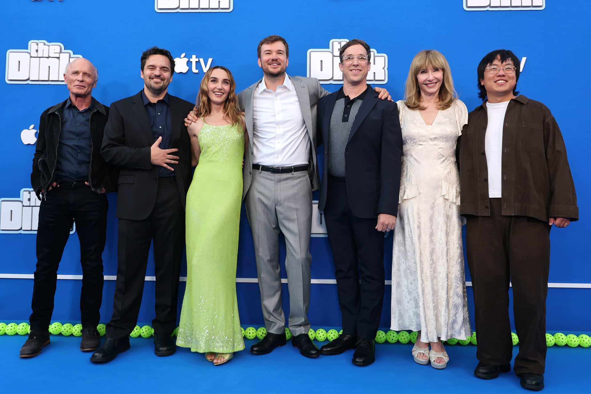 Seven people stand together smiling on a blue carpet in front of a blue backdrop with "Still" written on it, dressed in formal and semi-formal attire, posing for a group photo at The Dink premiere presented by Apple Original Films.