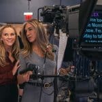 Two women stand together in a TV studio behind a camera and teleprompter, smiling and looking to the side. Studio crew and equipment are visible in the background, evoking the vibrant atmosphere of The Morning Show on Apple TV.