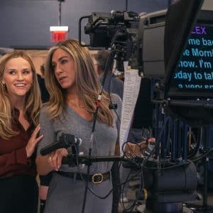 Two women stand together in a TV studio behind a camera and teleprompter, smiling and looking to the side. Studio crew and equipment are visible in the background, evoking the vibrant atmosphere of The Morning Show on Apple TV.