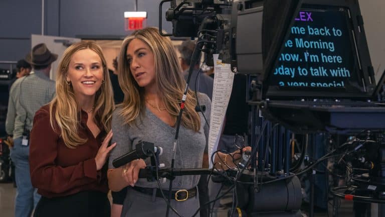 Two women stand together in a TV studio behind a camera and teleprompter, smiling and looking to the side. Studio crew and equipment are visible in the background, evoking the vibrant atmosphere of The Morning Show on Apple TV.