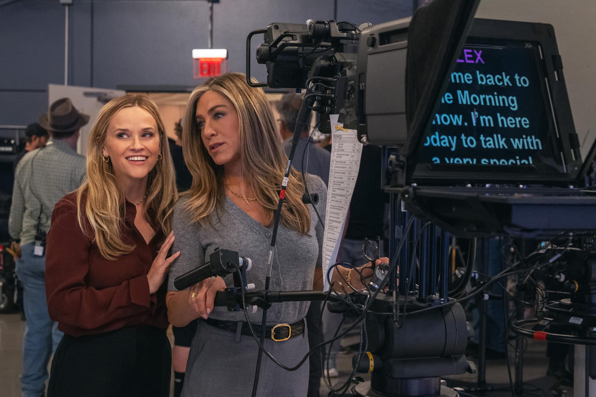 Two women stand together in a TV studio behind a camera and teleprompter, smiling and looking to the side. Studio crew and equipment are visible in the background, evoking the vibrant atmosphere of The Morning Show on Apple TV.