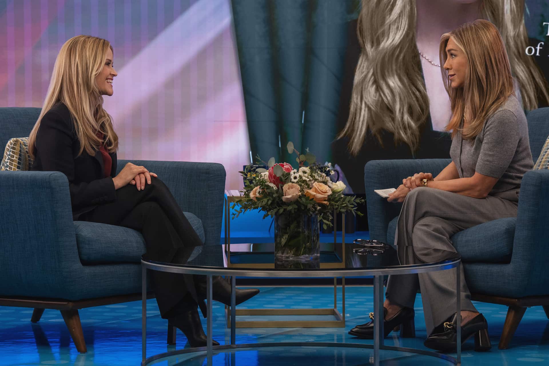 The Morning Show Season 5 - Two women sit across from each other on blue chairs, engaged in conversation on The Morning Show set. A bouquet of flowers rests on the glass table between them, and a large screen is visible in the background, capturing the lively atmosphere of Apple TV's acclaimed series.