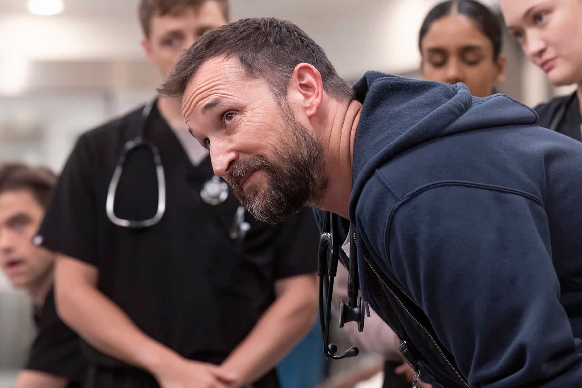 A bearded man in a navy hoodie, with a stethoscope around his neck, leans forward while a group of people in black scrubs watch attentively in a medical setting—reminiscent of the compelling scenes featured in Apple TV’s critically acclaimed series recognized with multiple Emmy nominations.