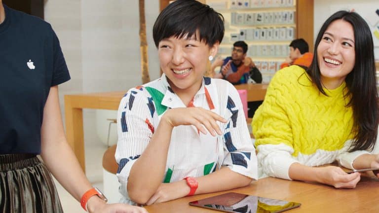 Three people sit at a wooden table in a brightly lit Apple Store, smiling and talking. One wears a patterned shirt, another a yellow sweater, and an iPad rests on the table as they discuss creative skills together.