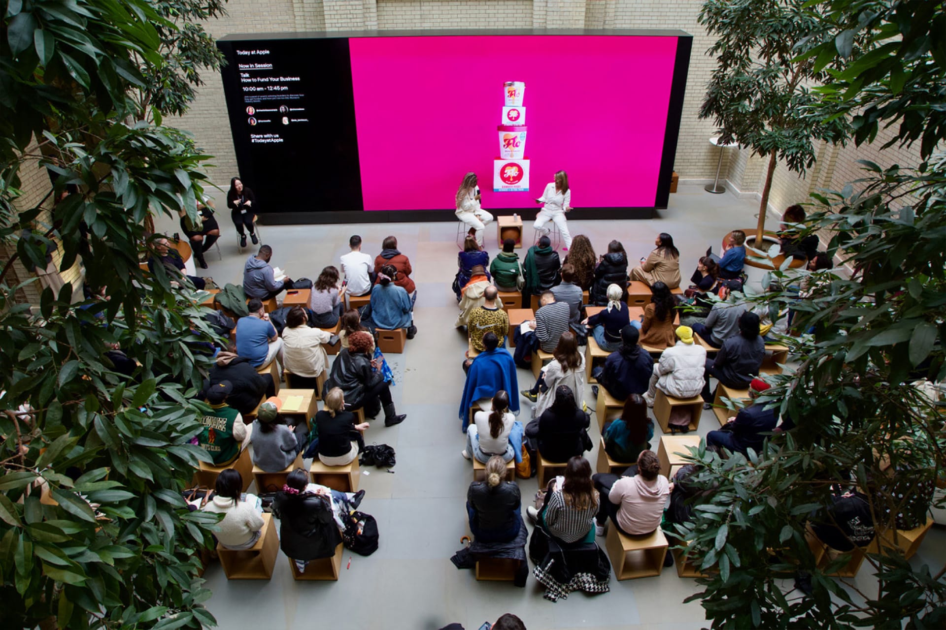 A large audience sits on wooden stools facing a bright pink screen, while three people on stage speak. The indoor event, part of Today at Apple, is surrounded by tall leafy trees and inspires guests to develop their creative skills.
