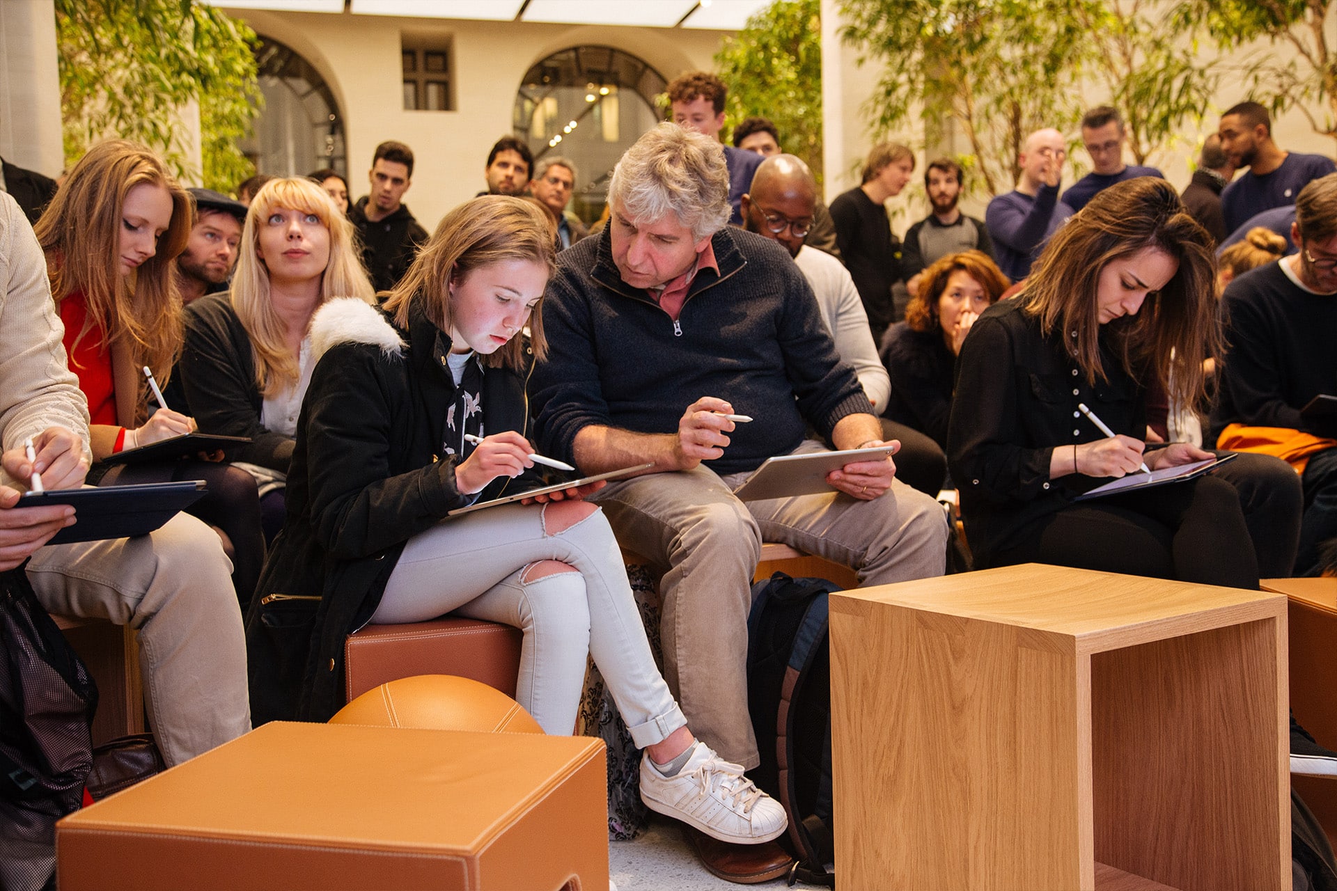 A group of people sits closely together on stools in a bright, modern space, focused on drawing or writing on tablets during a Today at Apple session. Trees and large windows are visible in the background.