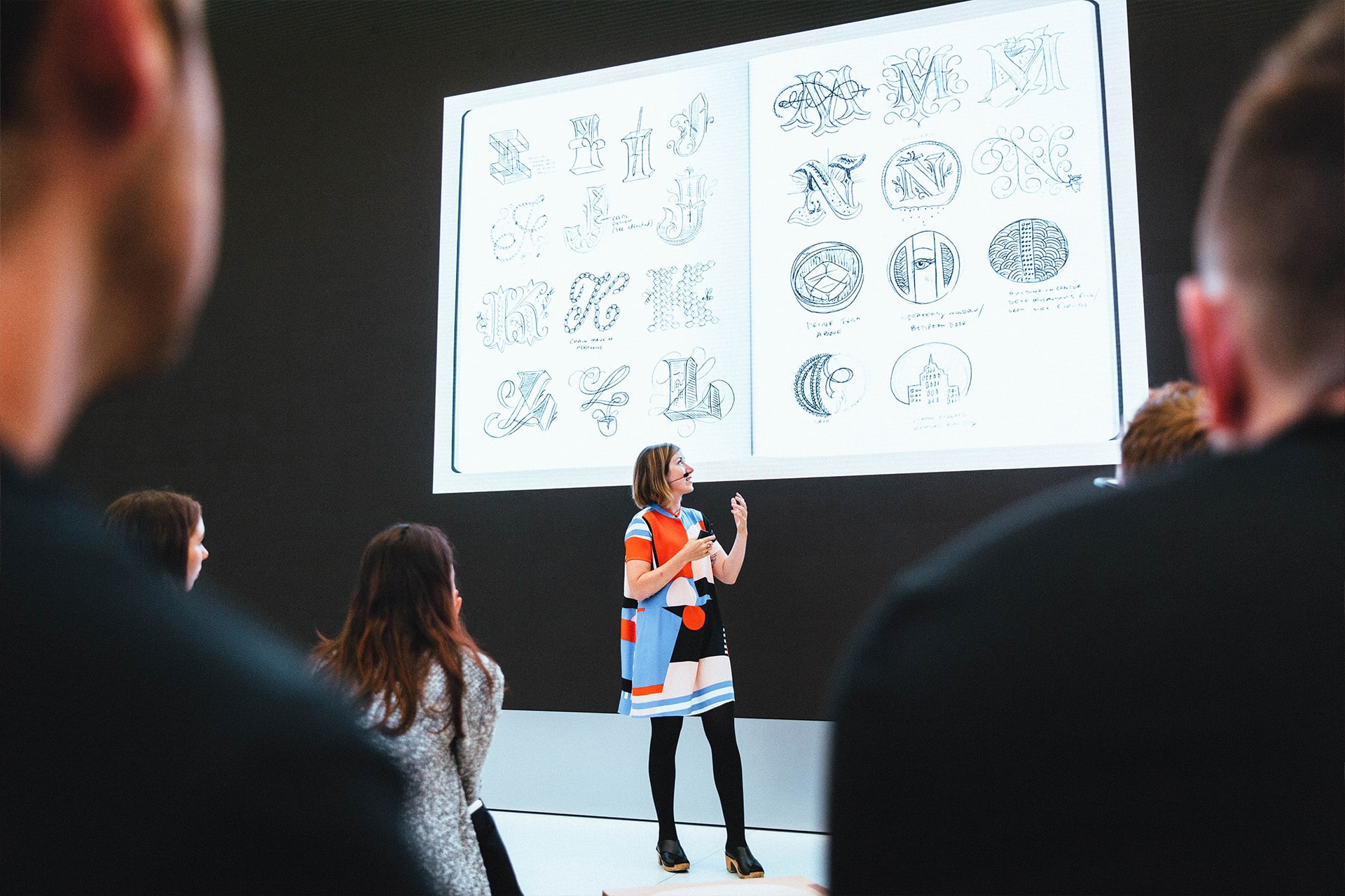 A woman in a colorful dress presents ornate letter designs on a large screen during an Apple Store education session, engaging the audience seated in front of her.