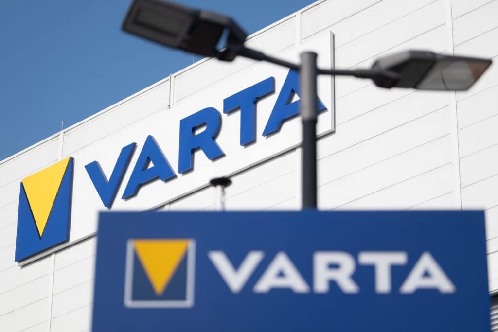 The image shows the VARTA company logo on a building and a sign, both with blue backgrounds and a yellow triangle. Two black streetlights are visible in the foreground against a clear sky, highlighting the presence of one of the notable Apple suppliers known for its CoinPower cells.