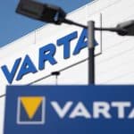 The image shows the VARTA company logo on a building and a sign, both with blue backgrounds and a yellow triangle. Two black streetlights are visible in the foreground against a clear sky, highlighting the presence of one of the notable Apple suppliers known for its CoinPower cells.