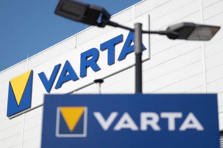 The image shows the VARTA company logo on a building and a sign, both with blue backgrounds and a yellow triangle. Two black streetlights are visible in the foreground against a clear sky, highlighting the presence of one of the notable Apple suppliers known for its CoinPower cells.