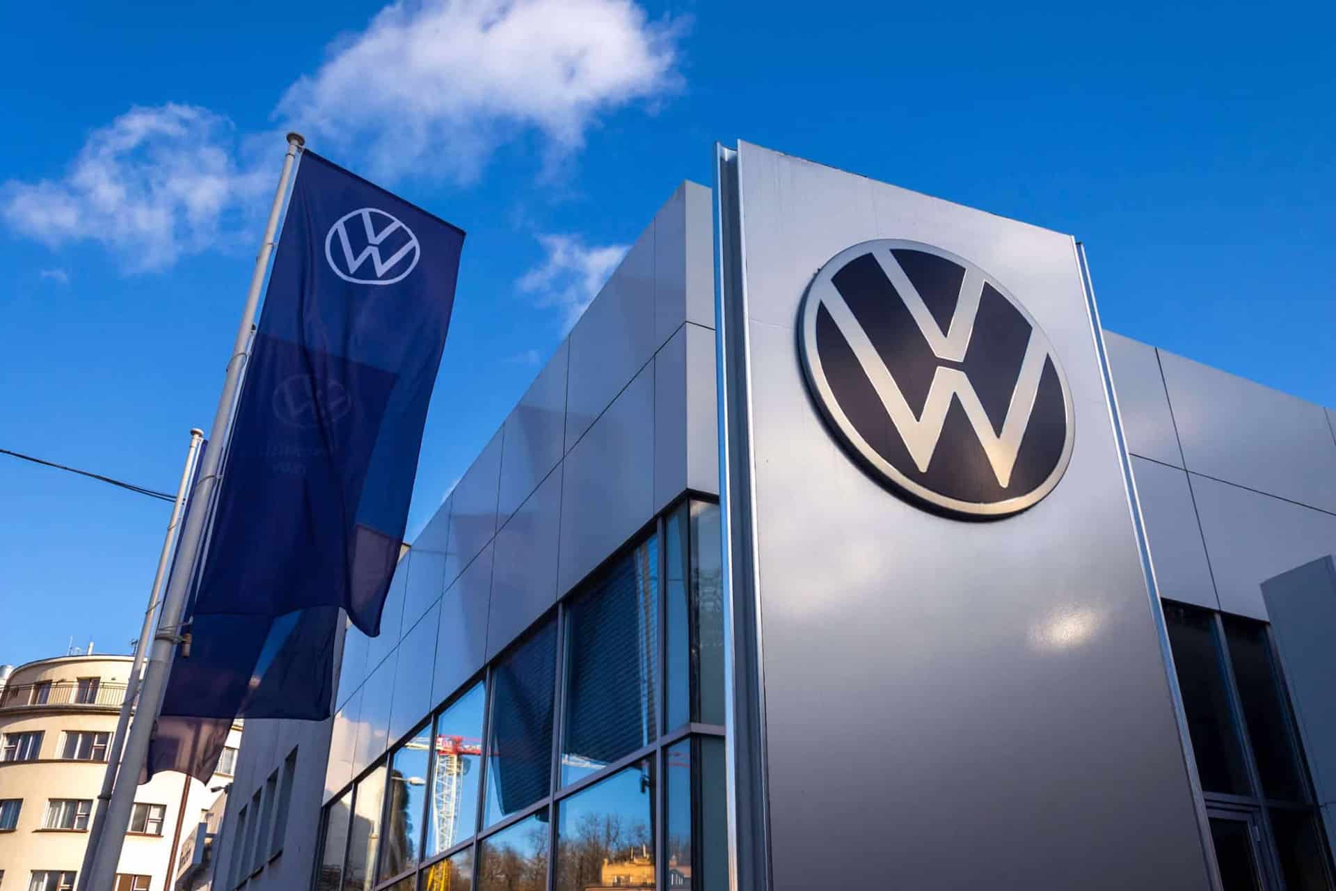 A Volkswagen dealership building with a large VW logo on the exterior wall and a VW flag, set against a blue sky with clouds—perfect for those interested in the latest Volkswagen Car Key features like Apple Wallet compatibility.