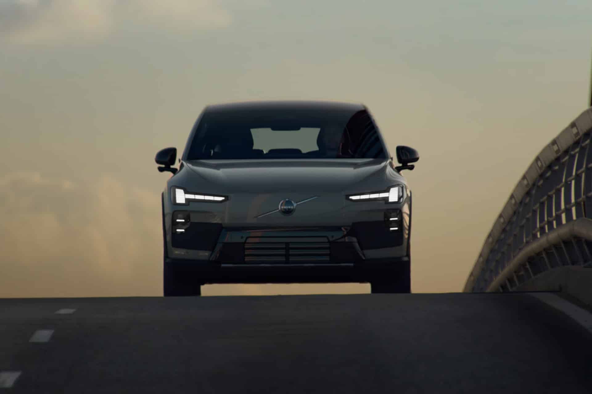 A modern gray car with sleek headlights drives up an empty road at sunset, viewed from the front against a softly lit sky and an urban bridge railing on the right, its advanced Volvo infotainment system seamlessly integrating Apple Music for a connected driving experience.