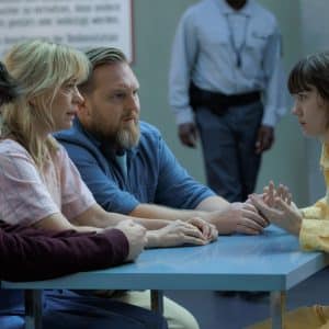 Four people sit at a table; three appear to be visitors, while one, wearing a yellow prison uniform, gestures while speaking. A guard stands in the background, and the setting resembles a visitation room featured in Where’s Wanda season two on Apple TV.