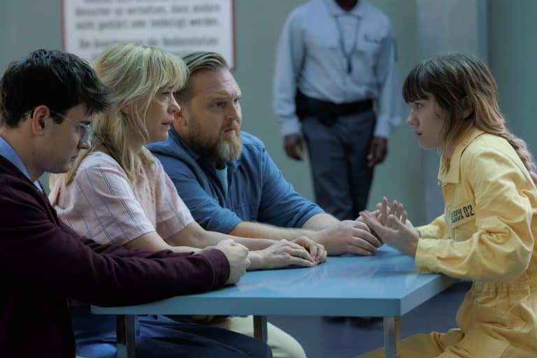 Four people sit at a table; three appear to be visitors, while one, wearing a yellow prison uniform, gestures while speaking. A guard stands in the background, and the setting resembles a visitation room featured in Where’s Wanda season two on Apple TV.