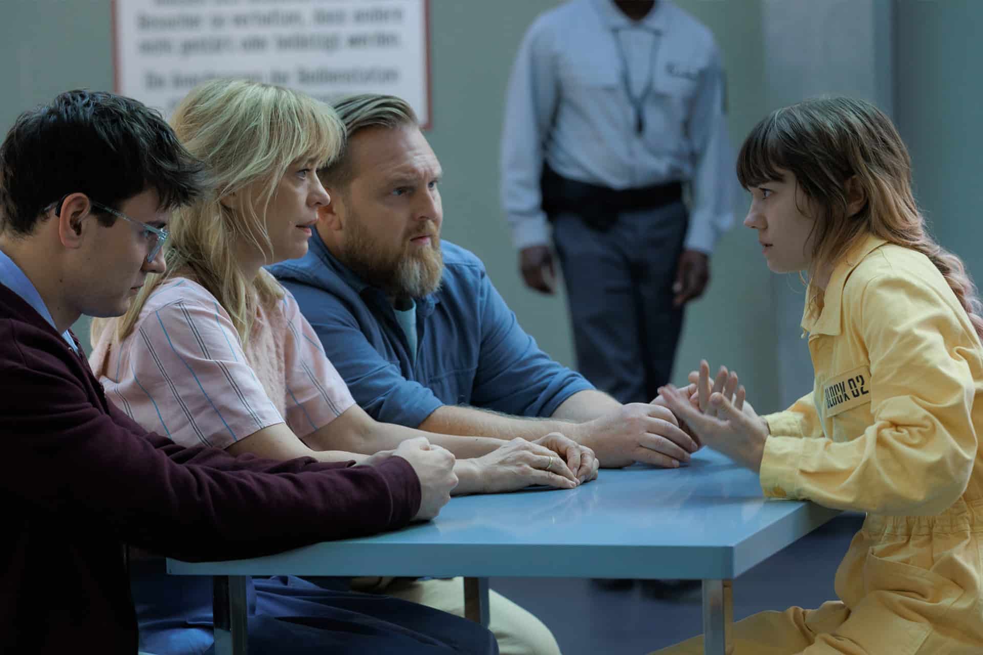 Four people sit at a table; three appear to be visitors, while one, wearing a yellow prison uniform, gestures while speaking. A guard stands in the background, and the setting resembles a visitation room featured in Where’s Wanda season two on Apple TV.