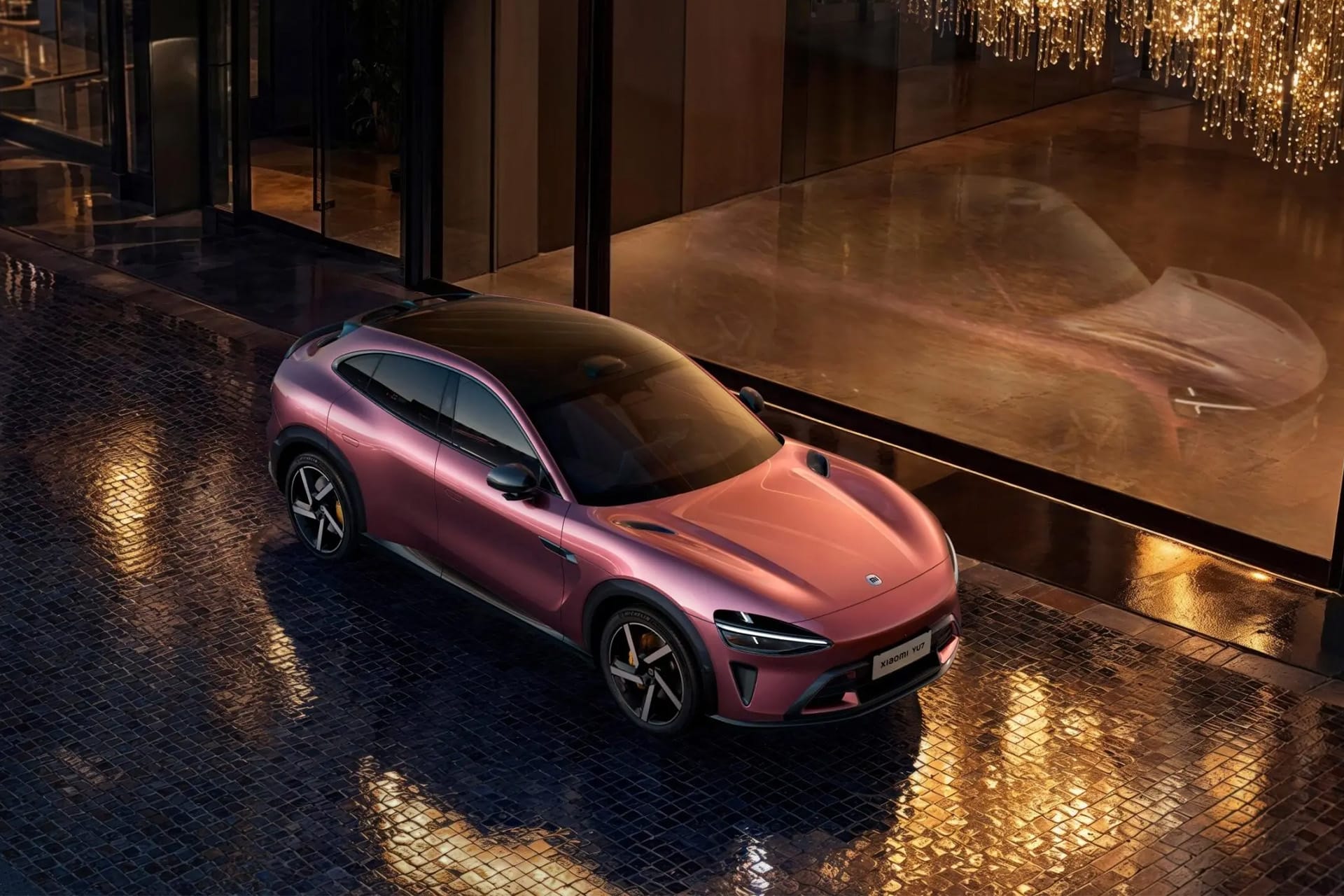 A sleek, pinkish-purple Lucid electric car is parked on wet cobblestone beside a glass building with golden chandeliers reflected on the surface, creating a luxurious nighttime atmosphere.