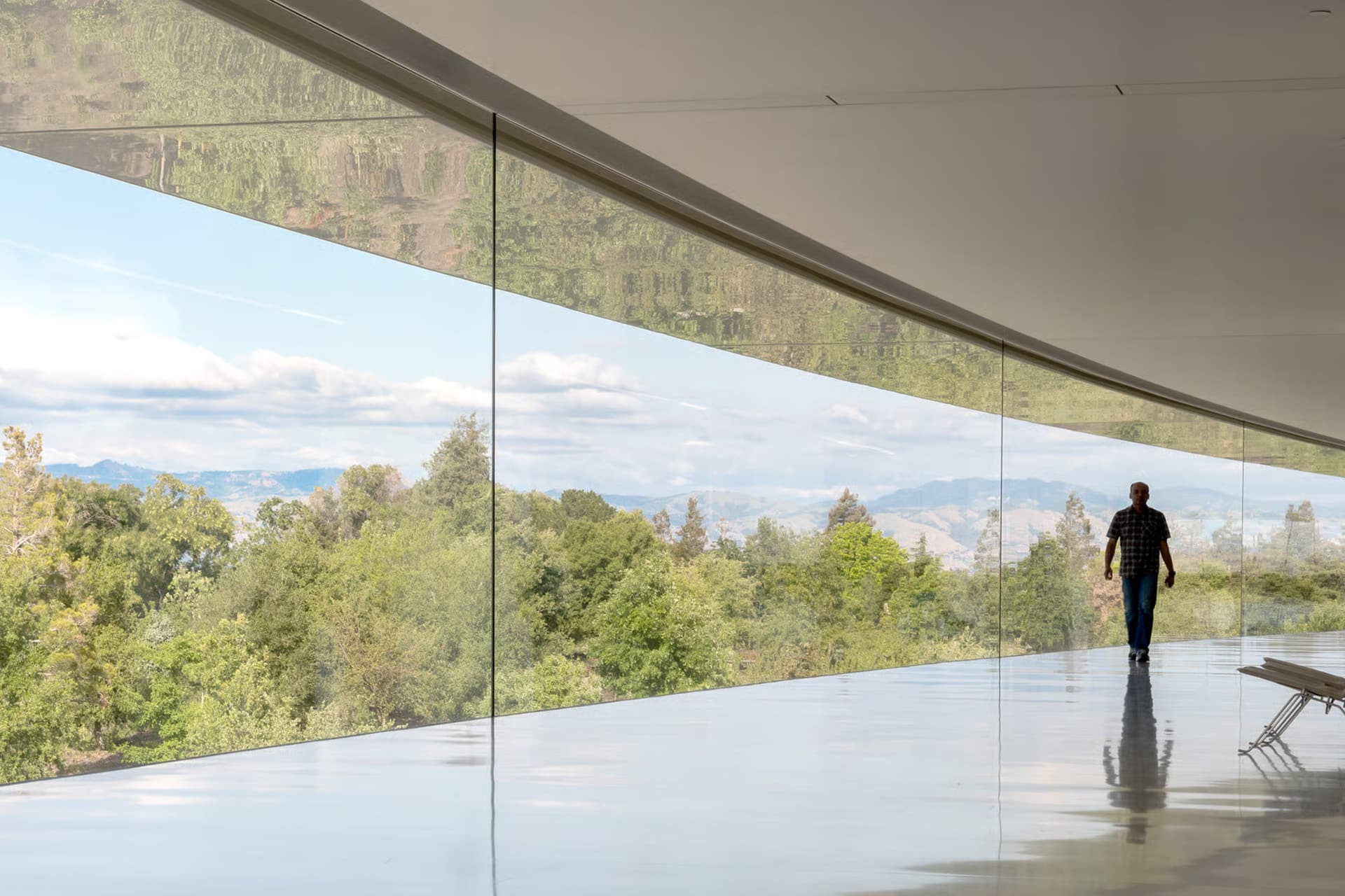 Apple Park details - A person walks along a modern building’s curved, glass-walled corridor, reminiscent of Apple Park architecture, with scenic views of trees and distant mountains under a partly cloudy sky.