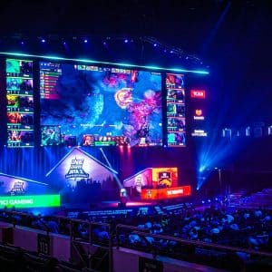 An esports arena with a large screen displays a video game match. Colored lights illuminate the stage, where teams play at desks. Spectators watch from the audience seats as excitement builds around every play. Team logos and sponsors are visible, reflecting the growing presence of modern betting platforms in the esports scene.