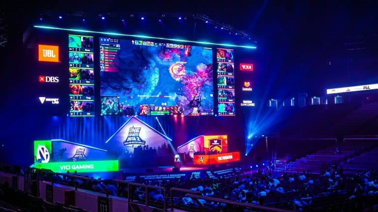 An esports arena with a large screen displays a video game match. Colored lights illuminate the stage, where teams play at desks. Spectators watch from the audience seats as excitement builds around every play. Team logos and sponsors are visible, reflecting the growing presence of modern betting platforms in the esports scene.