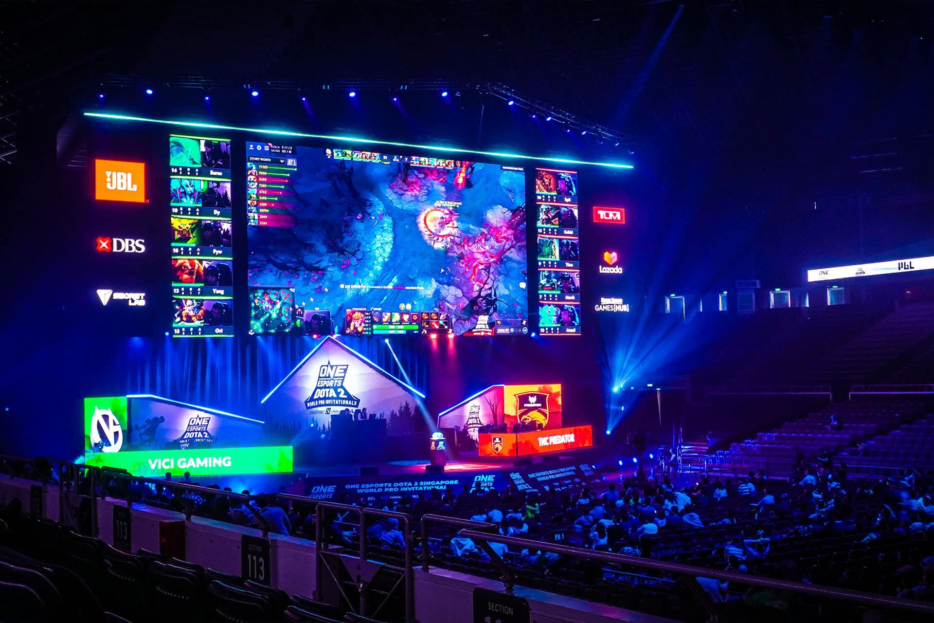 An esports arena with a large screen displays a video game match. Colored lights illuminate the stage, where teams play at desks. Spectators watch from the audience seats as excitement builds around every play. Team logos and sponsors are visible, reflecting the growing presence of modern betting platforms in the esports scene.