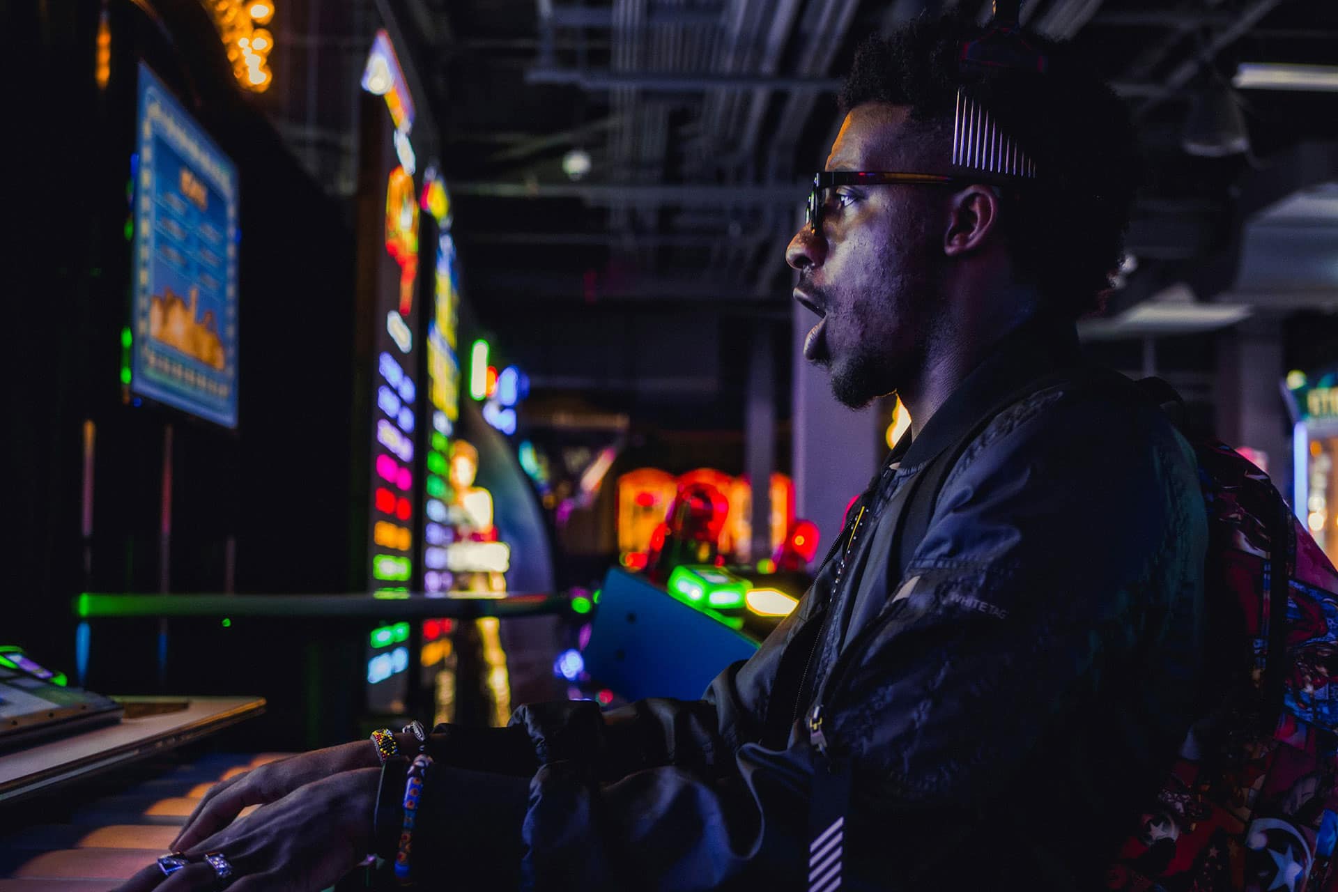 A person wearing glasses and headphones is playing an arcade game in a dimly lit room with colorful lights and game machines in the background, reminiscent of the dynamic atmosphere found in modern betting platforms.