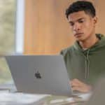 A young person in a green hoodie sits at a desk, working on a silver Apple laptop with a focused expression—perhaps researching the latest Apple security features or keeping up with recent Apple updates. The background is softly blurred and wooden.