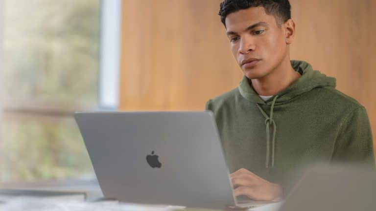 A young person in a green hoodie sits at a desk, working on a silver Apple laptop with a focused expression—perhaps researching the latest Apple security features or keeping up with recent Apple updates. The background is softly blurred and wooden.