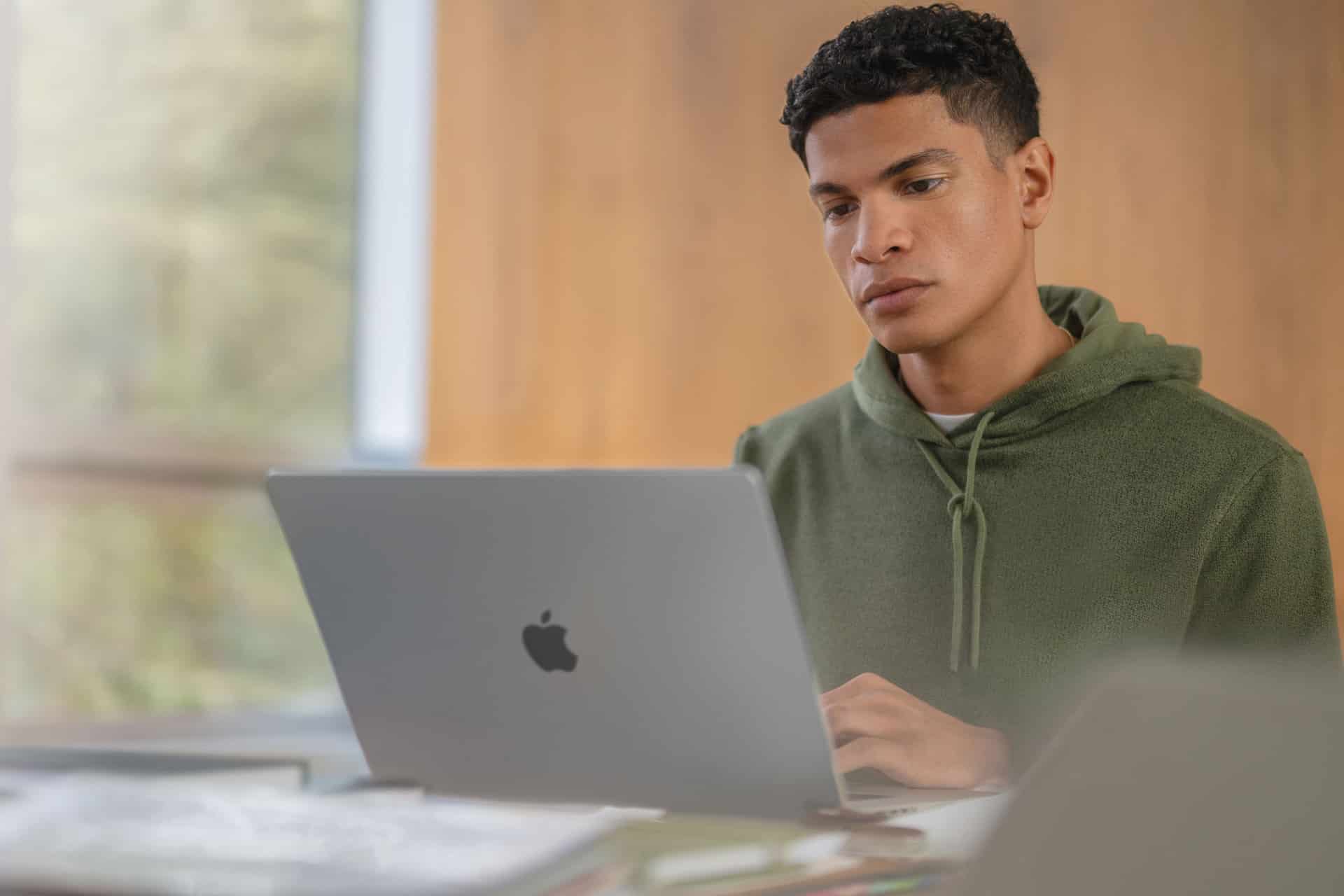 A young person in a green hoodie sits at a desk, working on a silver Apple laptop with a focused expression—perhaps researching the latest Apple security features or keeping up with recent Apple updates. The background is softly blurred and wooden.