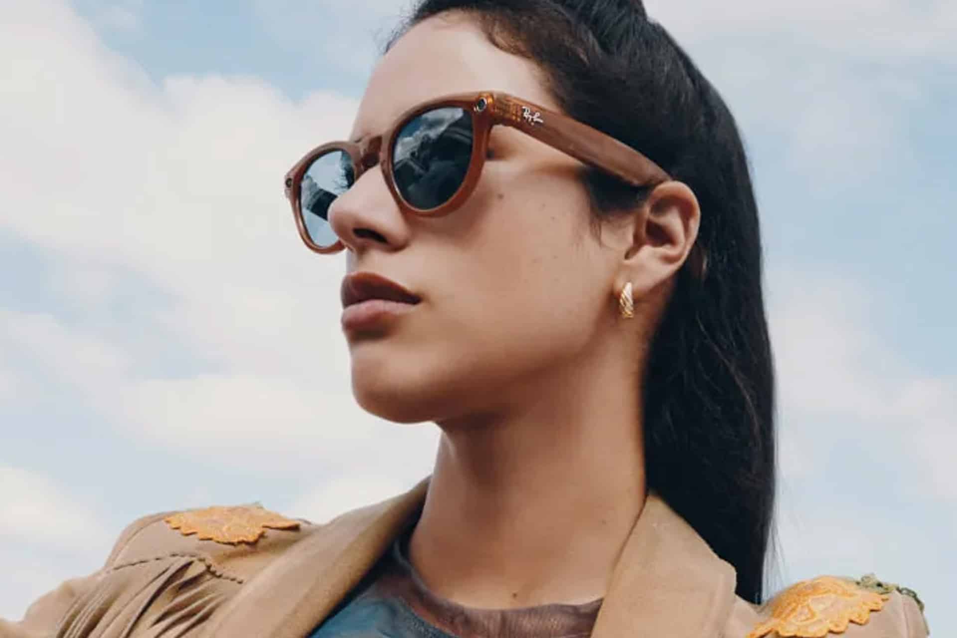 A woman wearing brown Ray-Ban Meta sunglasses and a tan jacket with yellow details looks off into the distance against a blue sky with clouds.
