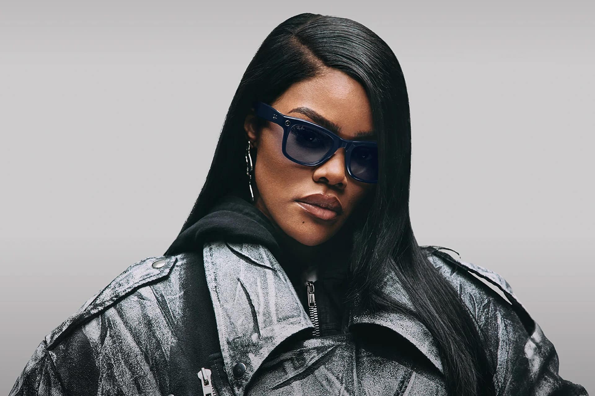 A woman with long straight hair wears Meta smart glasses, hoop earrings, a black hoodie, and a metallic silver jacket against a light grey background.