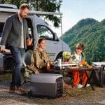 A man, woman, and child enjoy an outdoor meal at a campsite beside a camper van, surrounded by solar generators and mountains in the background, while their iPhone stays charged for capturing memories.