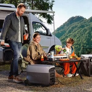 A man, woman, and child enjoy an outdoor meal at a campsite beside a camper van, surrounded by solar generators and mountains in the background, while their iPhone stays charged for capturing memories.