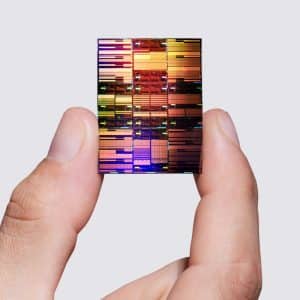 A hand holds a colorful, reflective 0.7nm chip between the thumb and index finger against a plain, light gray background.