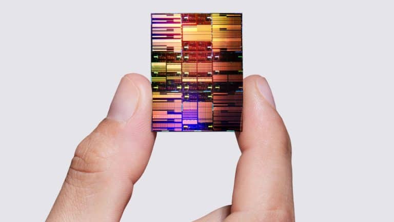 A hand holds a colorful, reflective 0.7nm chip between the thumb and index finger against a plain, light gray background.