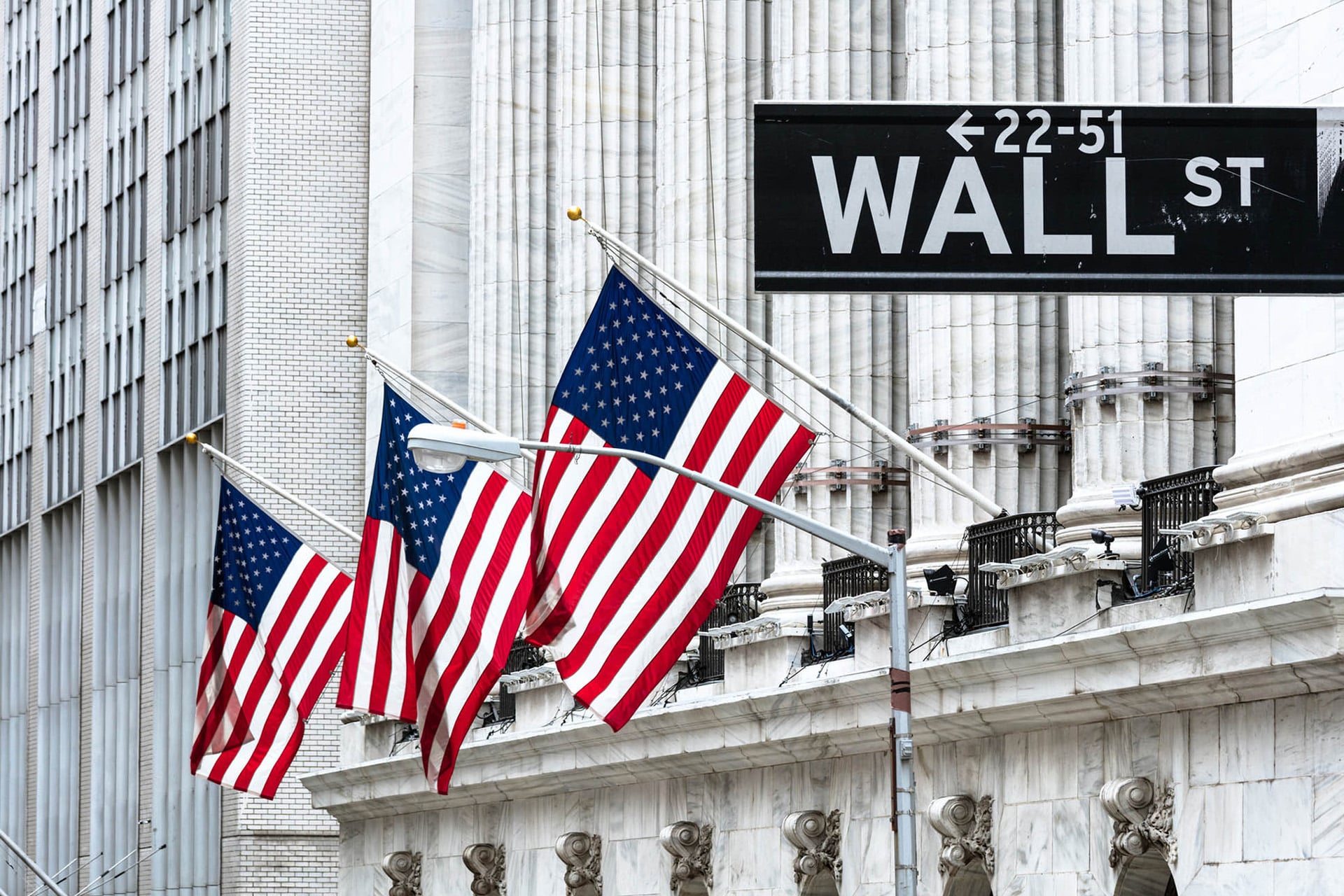 Three American flags hang in front of a building with tall columns and a street sign that reads “WALL ST” with directional arrows. The scene appears to be in a financial district, perhaps where news about Apple earnings and the latest Citi Apple target could shape the day’s trading on AAPL.
